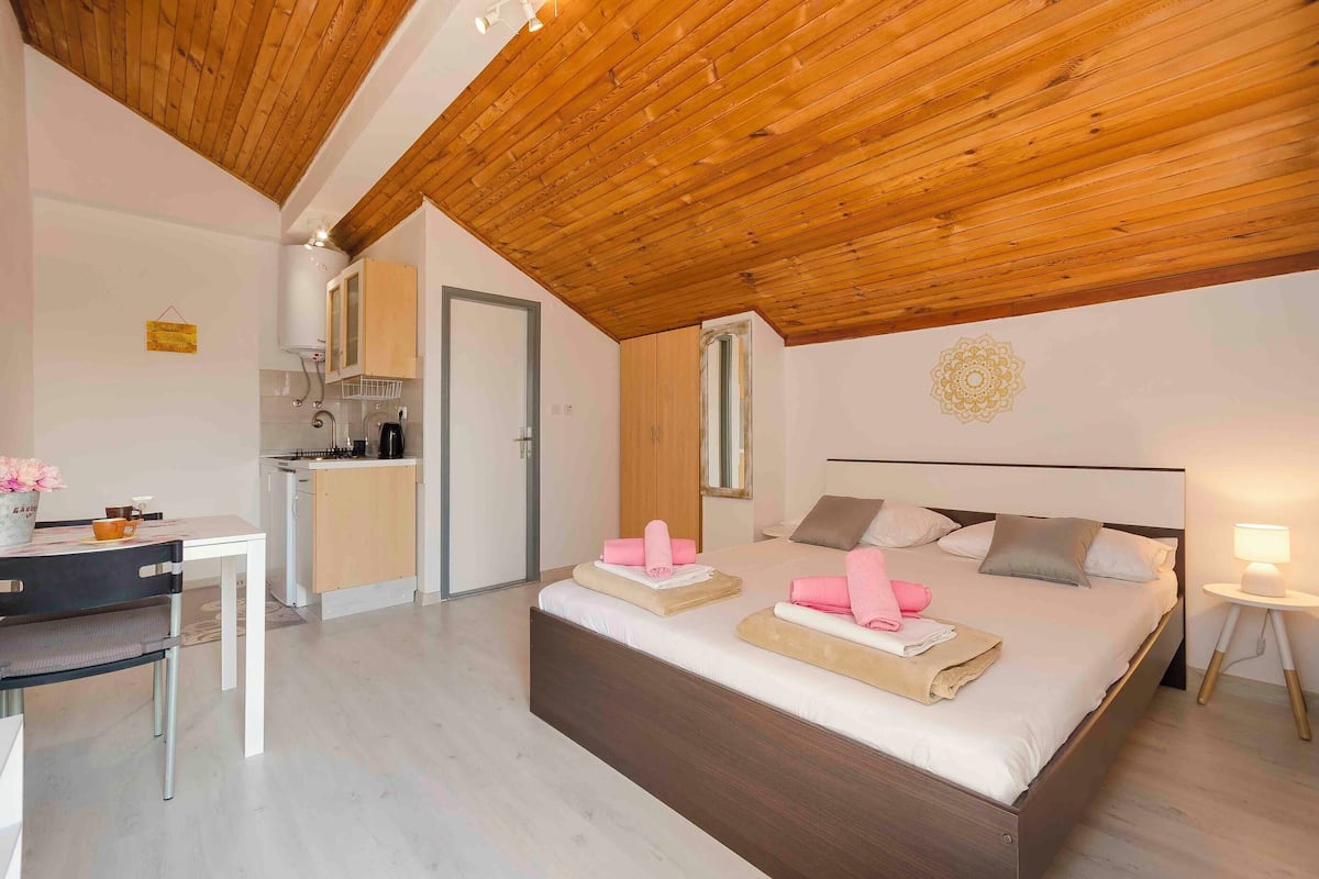 A modern bedroom with a wooden ceiling, a large bed with pink towels, a bedside table with a lamp, and a small kitchenette in the background.