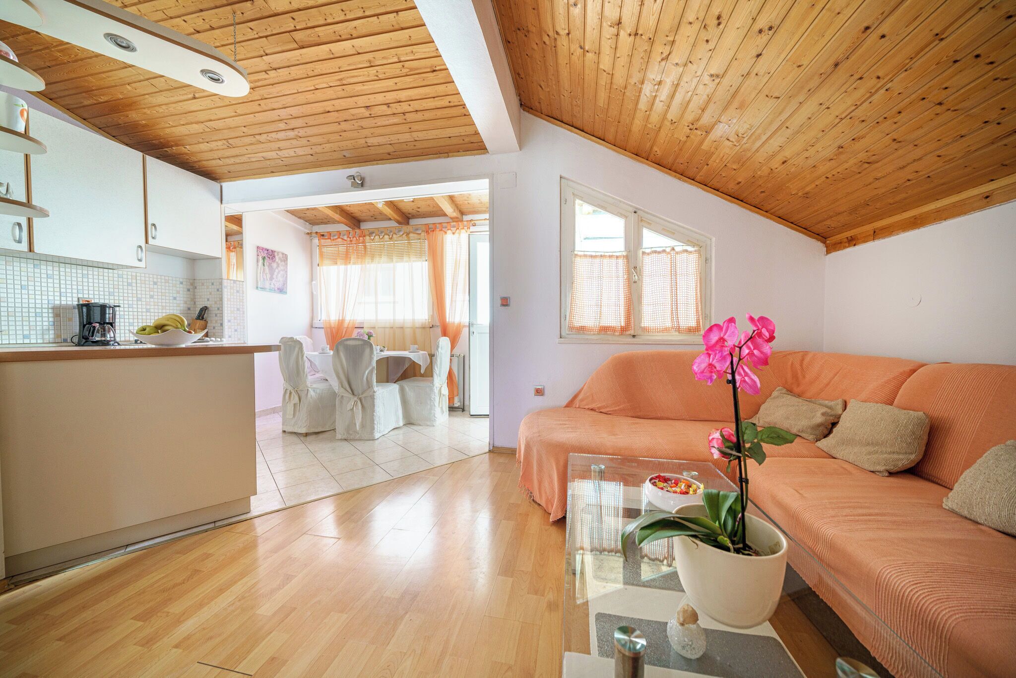 A spacious living area with a wooden ceiling, a sofa, a glass coffee table, and a kitchen area with a dining set.