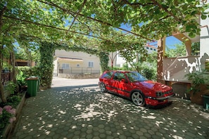 A red car parked under a tree in a paved courtyard with a building in the background. - Apartments Ivka (Vodice)