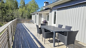 Eine Holzterrasse mit Tisch und Stühlen, ein überdachter Außenbereich und ein weißes Gebäude mit vertikaler Schalung.