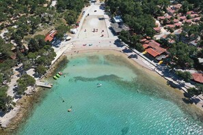An aerial view of a coastal area with clear turquoise waters, a sandy beach, and a row of buildings with red roofs. - Apartment Jadranka (Biograd na Moru)