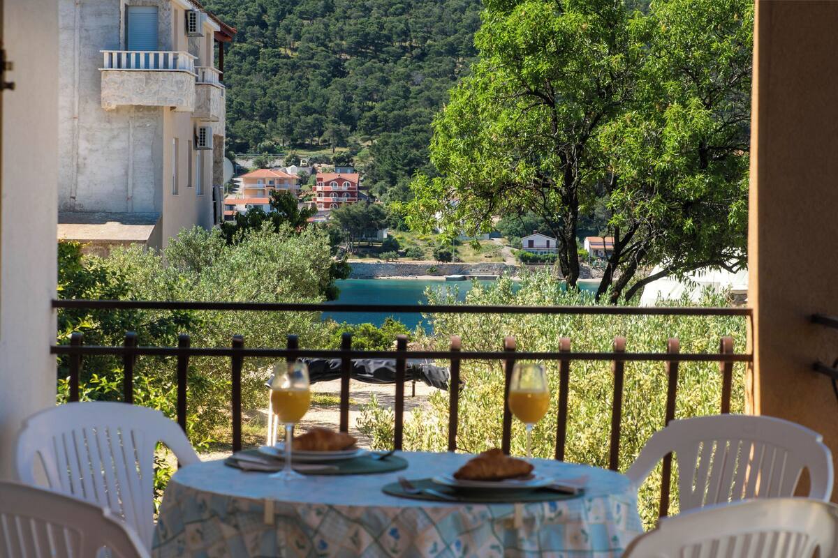 A balcony with a table set for breakfast, overlooking a scenic view of a coastal town and hills.