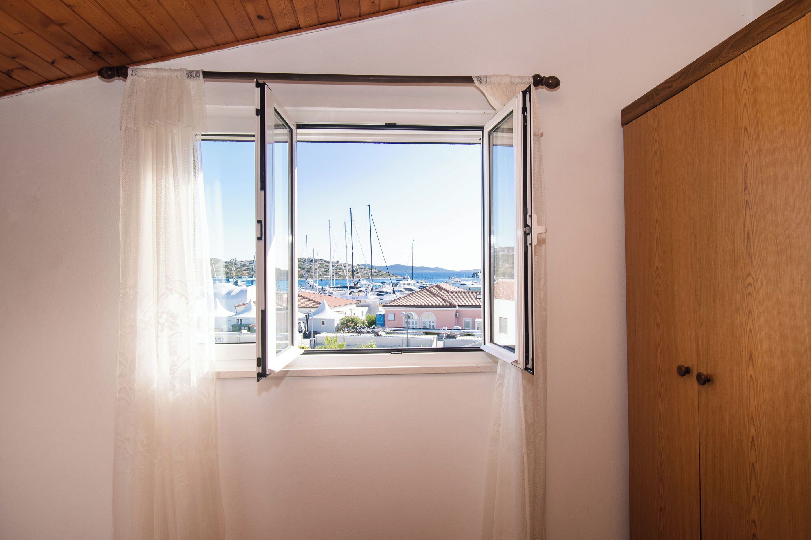 A room with a wooden door and a window with white curtains, offering a view of boats and buildings.