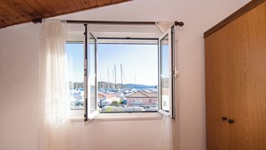 A room with a wooden door and a window with white curtains, offering a view of boats and buildings. - Apartments Nena (Tribunj)