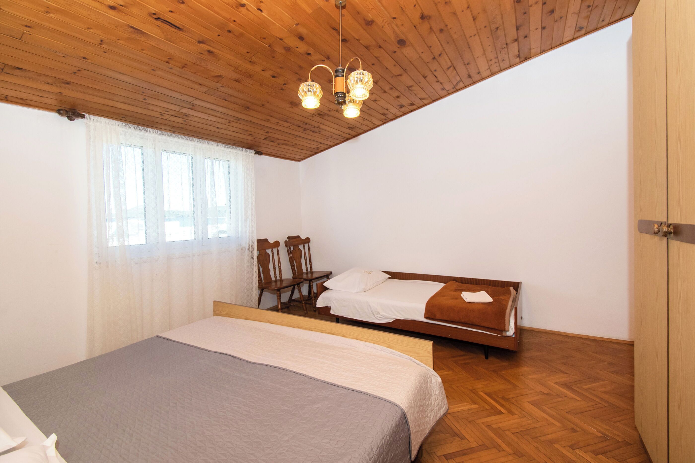 A bedroom with a bed, a wooden ceiling, a window with curtains, a chair, and a bedside table.