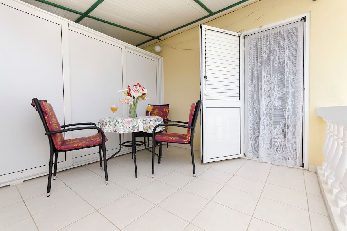 A balcony with a table and chairs, a vase with flowers, and a white door with a lace curtain.