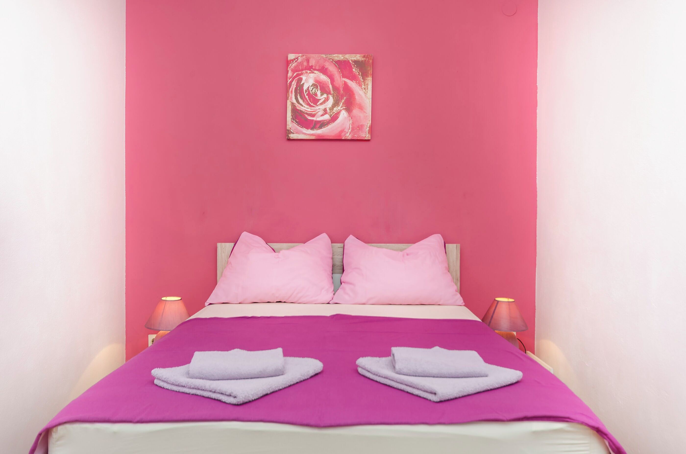 A hotel room with a bed, pink pillows, a purple blanket, and a pink wall.