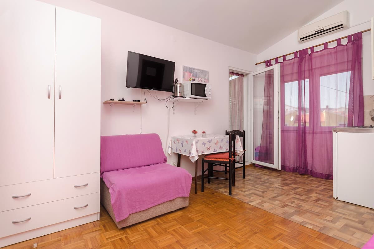 A small room with a bed, a table with a chair, a TV, a microwave, and a window with curtains.
