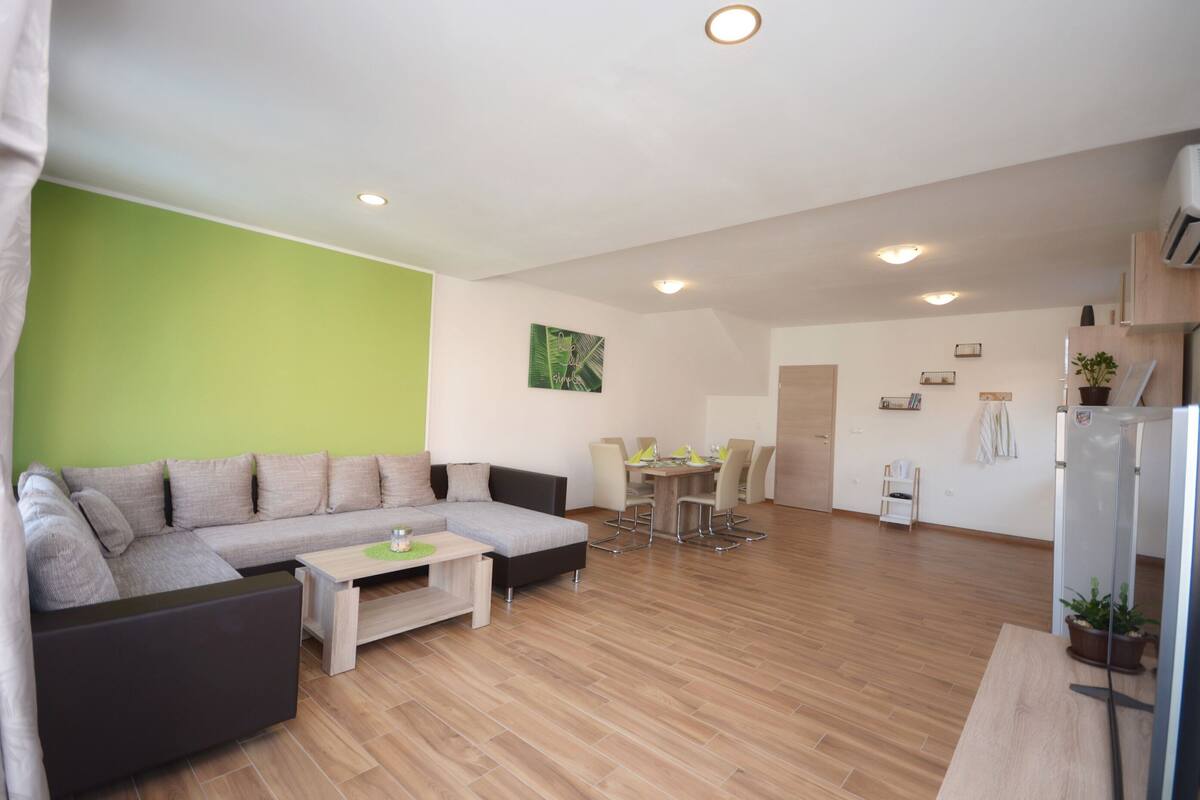 A modern living room with a sectional sofa, a dining table with chairs, and a small kitchen area with a refrigerator.