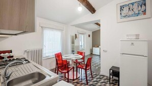 Cocina compacta con estufa a gas, pileta y comedor con sillas rojas. Se observa un refrigerador en una esquina.