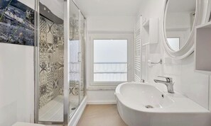 Une salle de bain moderne dotée d’une grande baignoire, d’une douche à parois de verre et offrant une vue sur la mer par une fenêtre.
