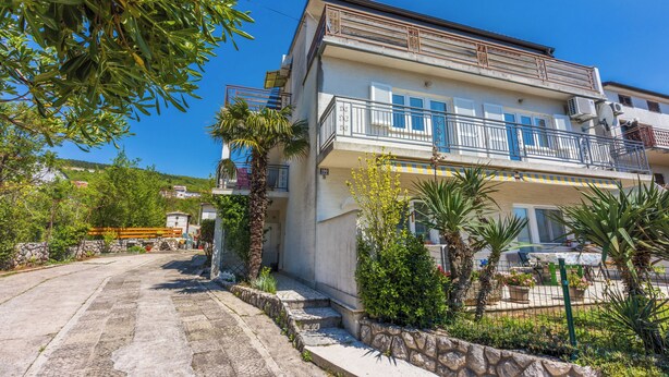 Un edificio de varios pisos con balcones, un sendero de piedra y vegetación circundante.