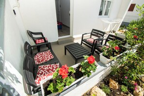 A patio with two black chairs, a small table, and a bench. There are potted plants with red flowers and a white railing. - Apartments Lily (Privlaka)