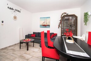 A modern living room with a grey sofa, red chairs, a dining table, a clock, a sailboat painting, and a cabinet with glass doors. - Apartments Lily (Privlaka)
