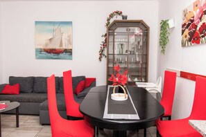 A modern living room with a round black table, red chairs, a grey sofa with red cushions, a glass cabinet with decorative items, and a sailboat painting on the wall. - Apartments Lily (Privlaka)