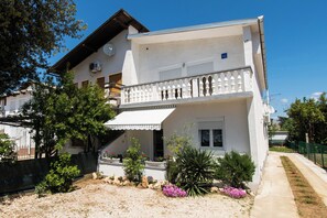 A two-story building with a balcony, a covered entrance, and a garden with various plants and flowers. - Apartments Lily (Privlaka)