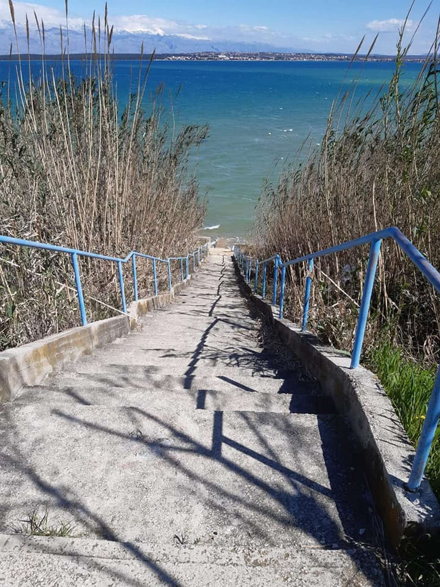 A concrete staircase leading to a beach with a clear view of the sea and distant mountains.