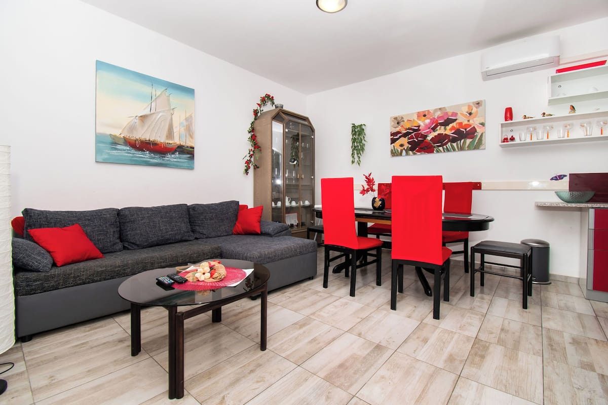 A modern living room with a grey sofa, red cushions, a coffee table with remote controls, a dining area with red chairs, and a kitchen with a white countertop.
