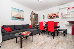 A modern living room with a grey sofa, red cushions, a coffee table with remote controls, a dining area with red chairs, and a kitchen with a white countertop. - Apartments Lily (Privlaka)