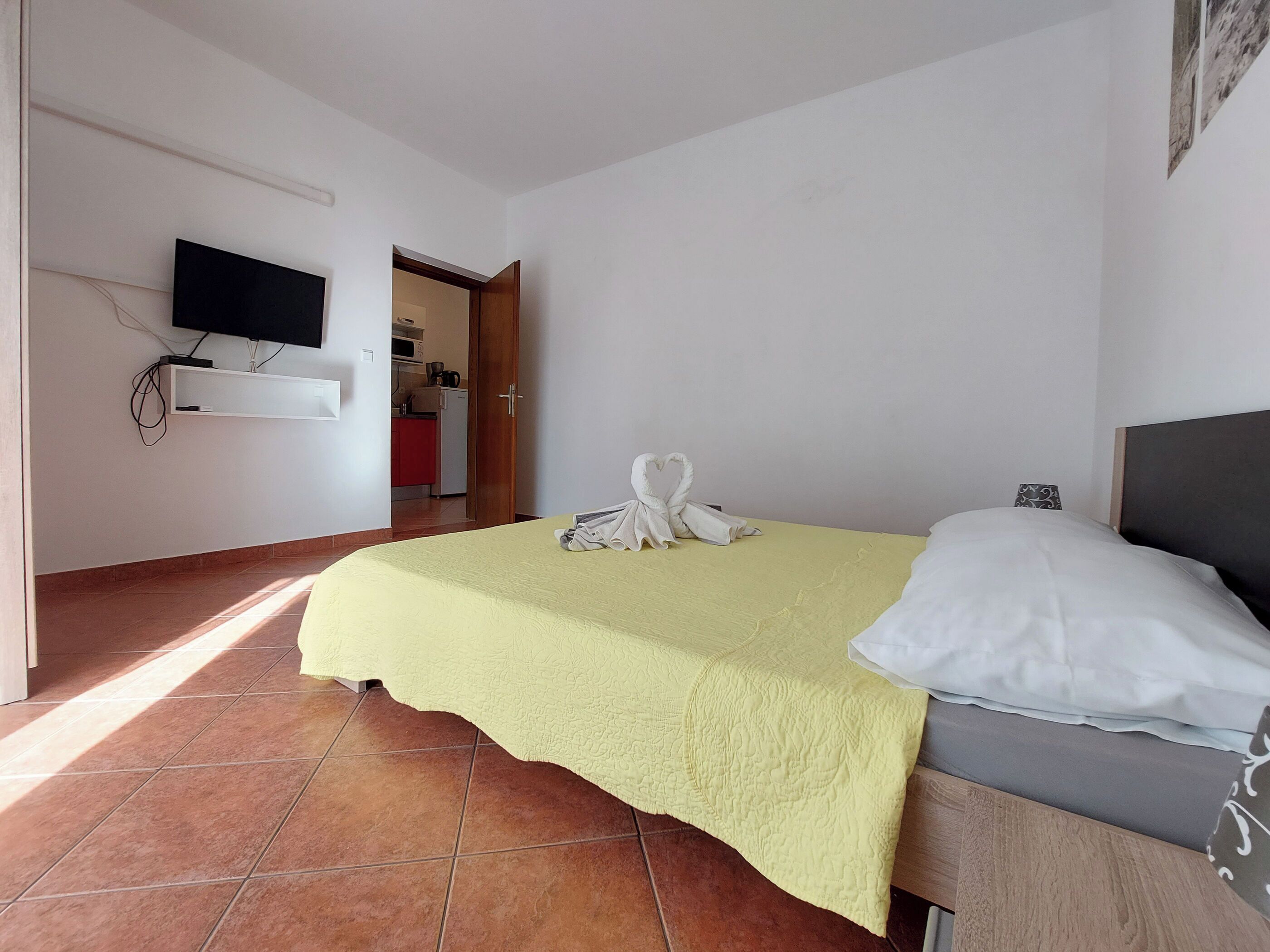 Habitación de hotel con cama, televisor montado en la pared y una cocina visible a través de una puerta abierta.