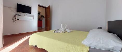 Habitación de hotel con cama, televisor montado en la pared y una cocina visible a través de una puerta abierta.