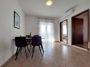 A dining area with a table and chairs, a framed picture on the wall, and a view through a window with sheer curtains. - Apartments Nono (Crikvenica)