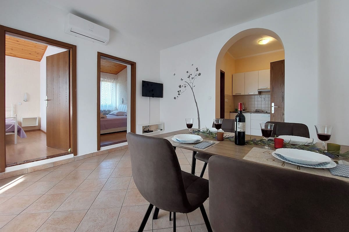Zona pranzo con tavolo apparecchiato per due, TV a parete e cucina visibile tramite un arco.