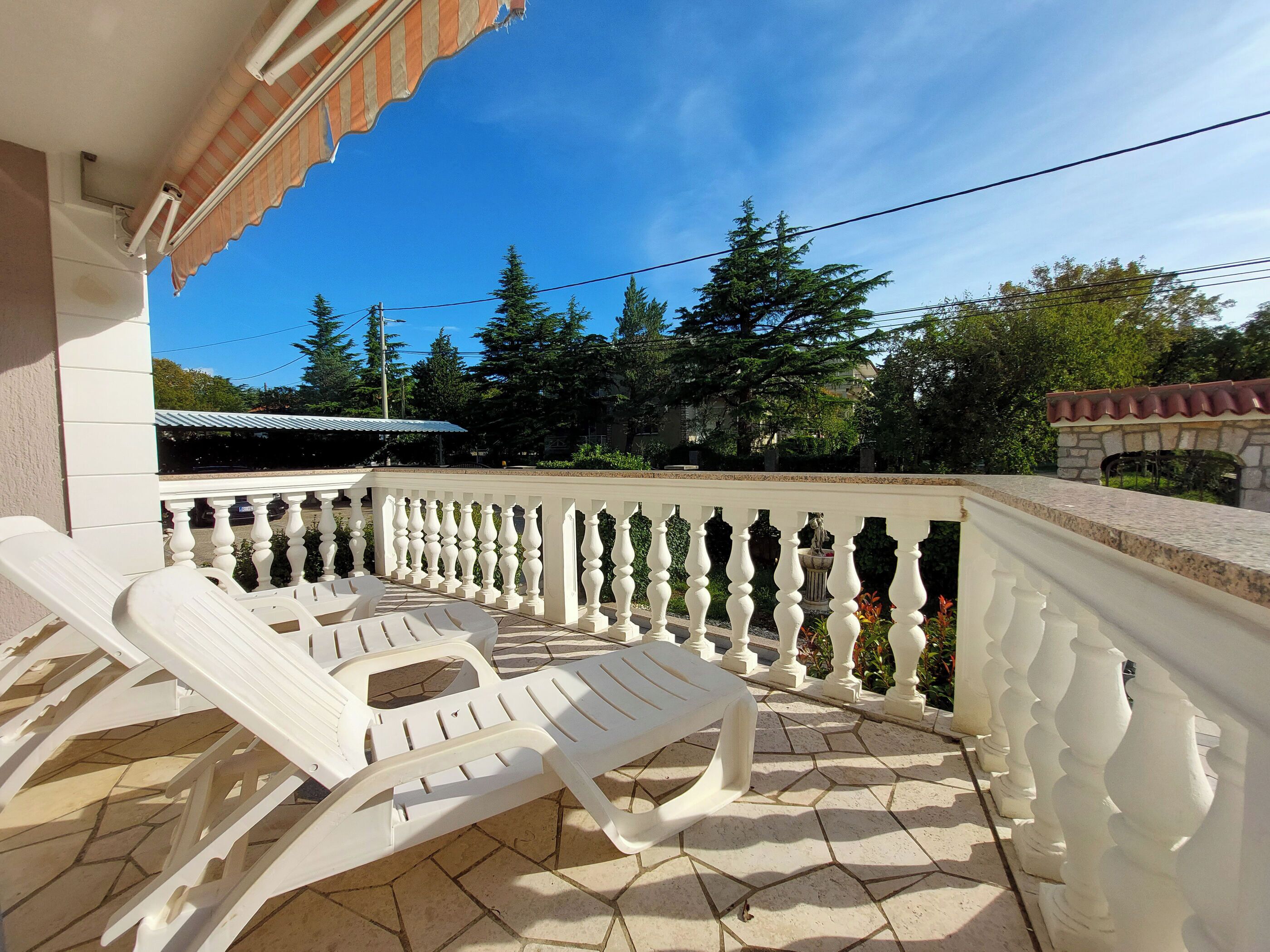 A balcony with white plastic chairs and a tiled floor, overlooking a garden with trees and a stone wall.