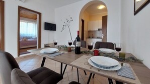 Spiseplads med et bord dækket til fire personer, en vinflaske og to glas rødvin. Der er et tv monteret på væggen, og et lille køkken er synligt gennem en bue.