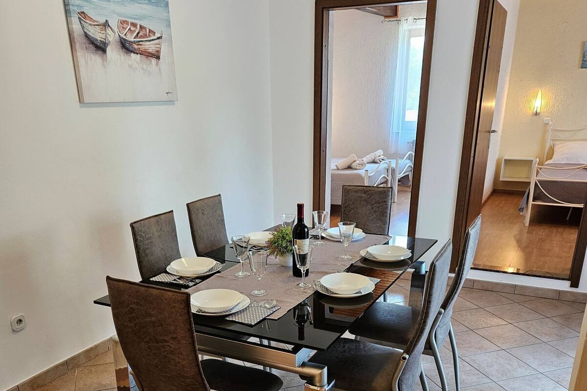 Un'area pranzo con un tavolo apparecchiato per quattro persone, una bottiglia di vino e un quadro raffigurante barche al muro.