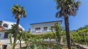 A two-story building with a white facade and blue trim, surrounded by a garden with palm trees and a variety of plants. - Apartments Dusanka (Crikvenica)