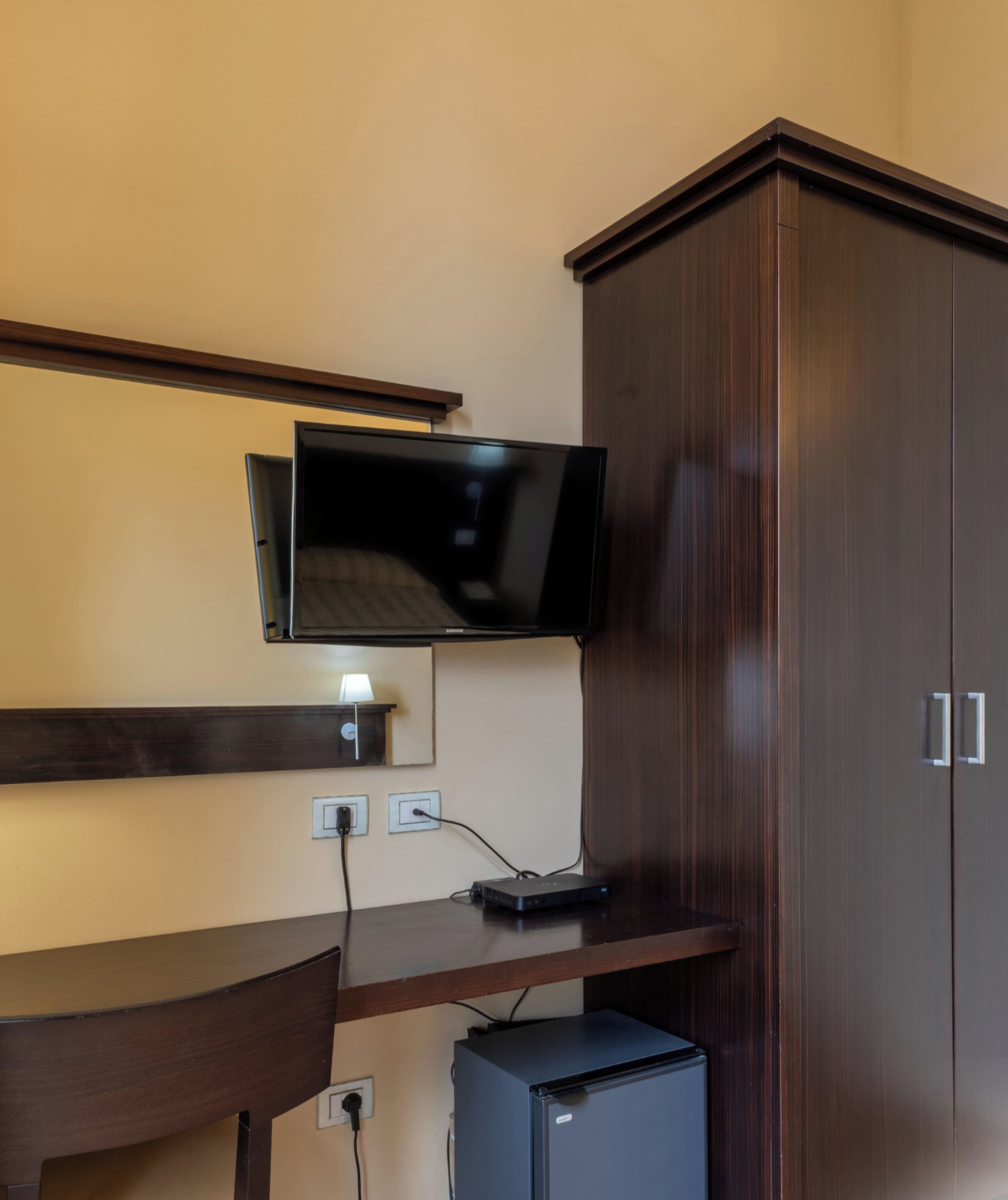 A room with a flat-screen TV mounted on the wall, a wooden desk with a computer, and a small refrigerator.