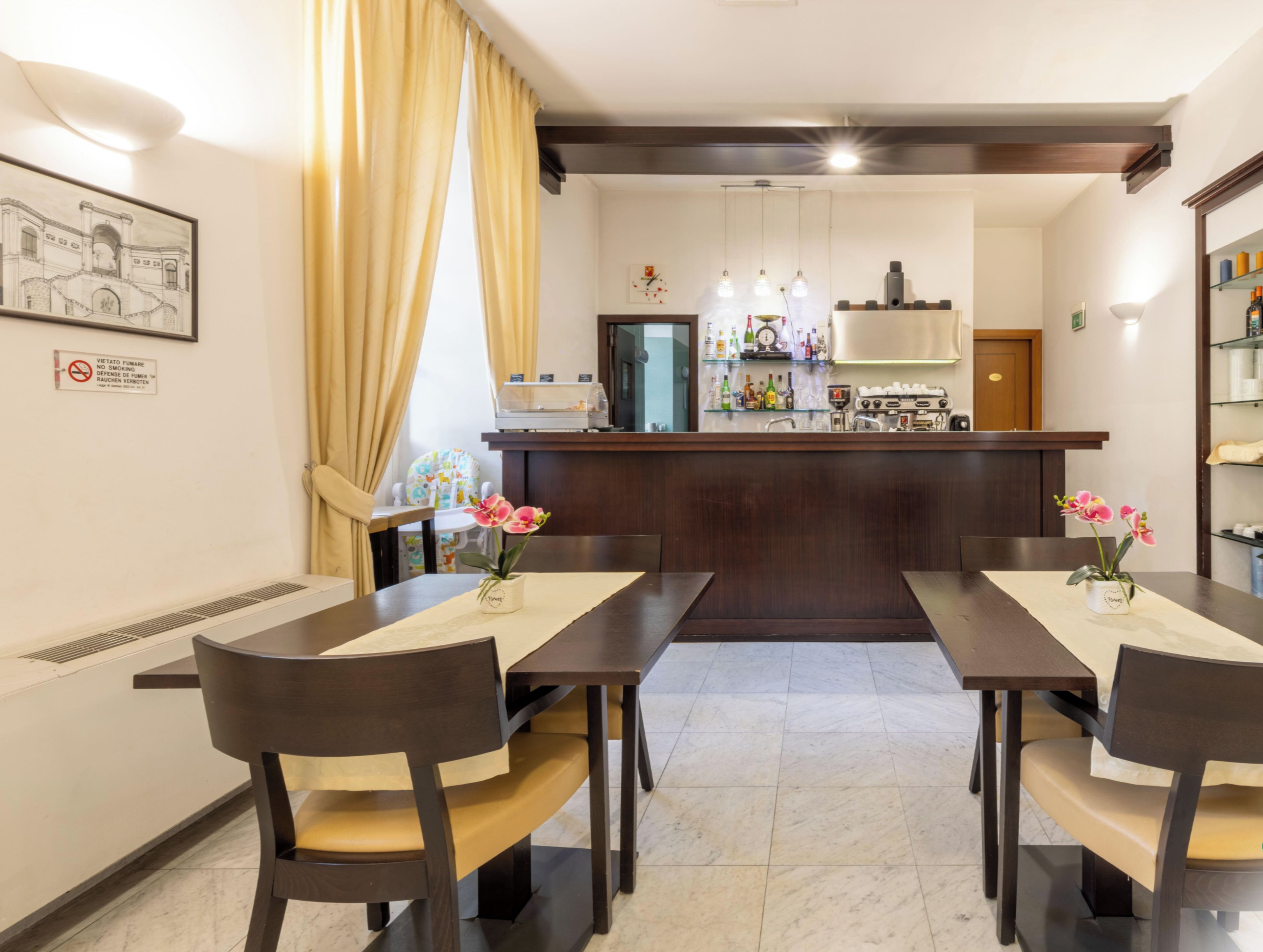 A hotel dining area with a bar in the center, two chairs at each end, and a table with a vase of flowers.