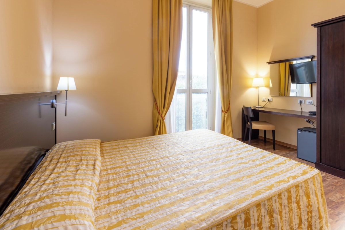 Ein Hotelzimmer mit Bett, Schreibtisch, Stuhl, Kleiderschrank und einem fenster mit Vorhängen.