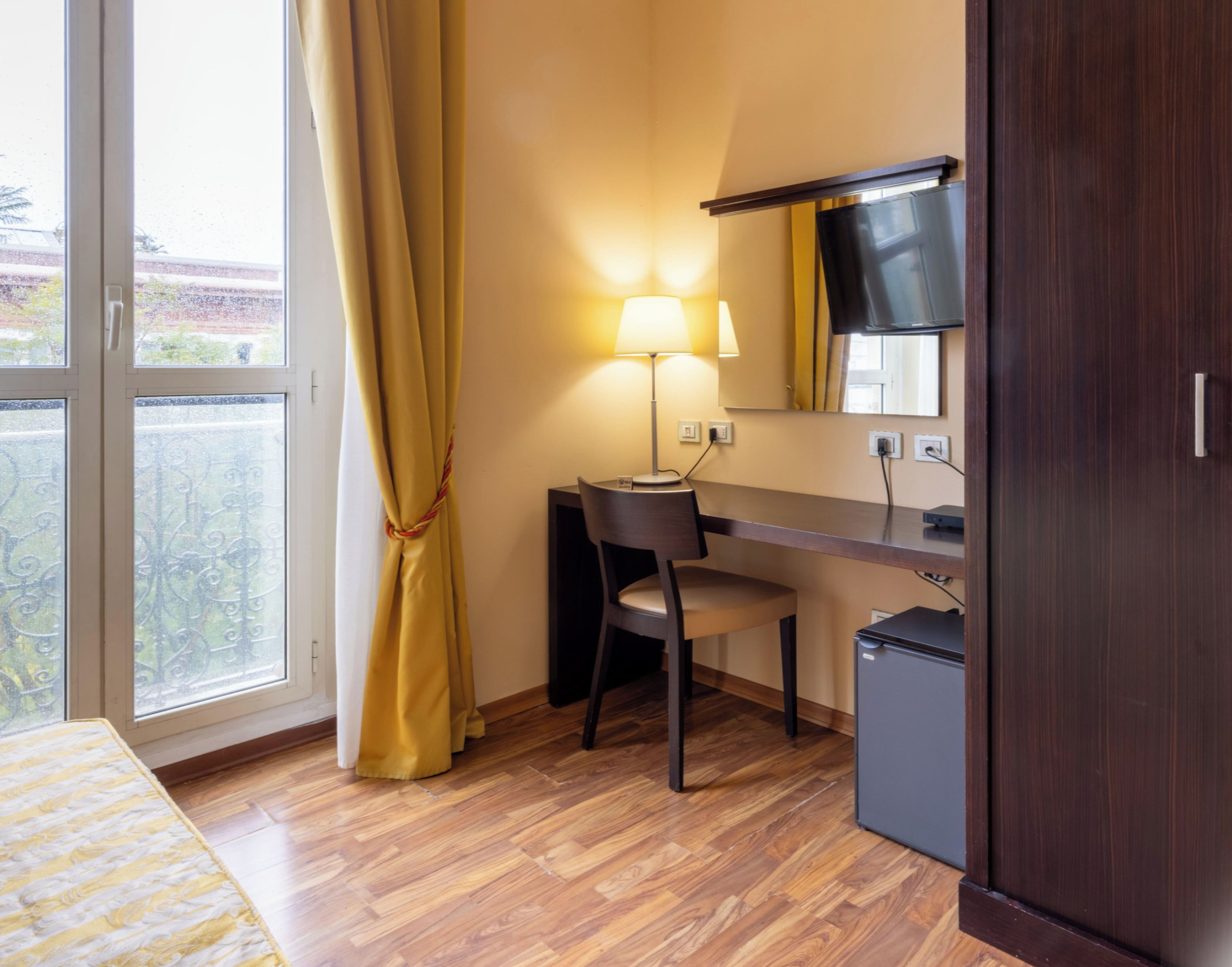 Habitación de hotel con piso de madera, un escritorio con silla, un refrigerador pequeño, una cama con colcha a rayas y una ventana con cortinas.