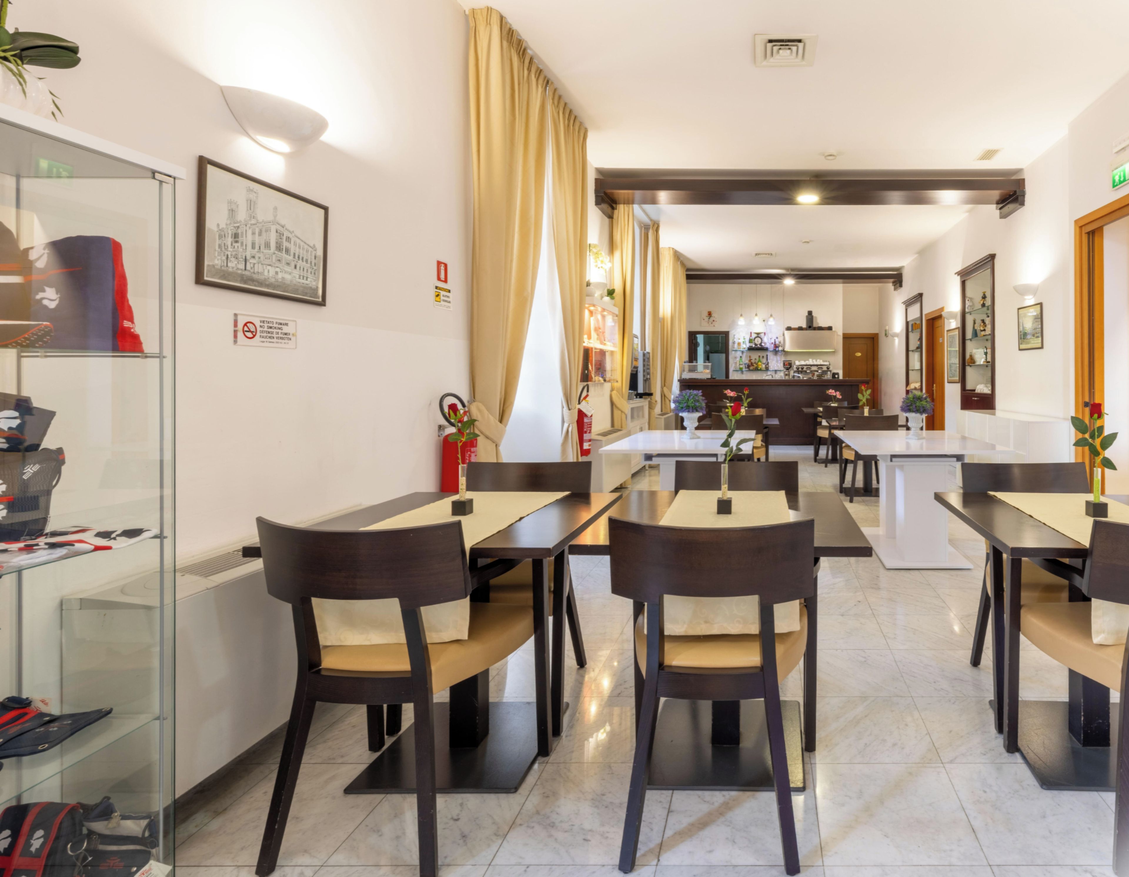 A hotel dining area with multiple tables and chairs, a kitchen in the background, and a glass display case with items.