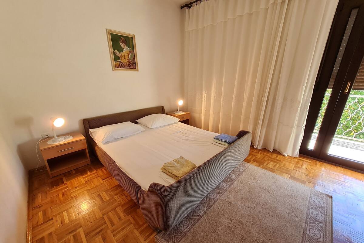 A bedroom with a large bed, two bedside tables, a nightstand, a painting on the wall, and a view through the window.