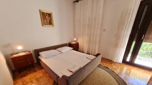 A bedroom with a wooden floor, a bed with white linens, two bedside tables with lamps, a painting on the wall, and a window with curtains. - Apartment Andrea (Novi Vinodolski)