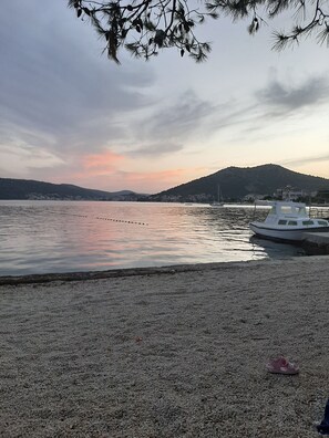 Ein Kiesstrand mit einem Boot am rechten Rand, eine ferne Küstenlinie mit Gebäuden und ein Berg im Hintergrund.