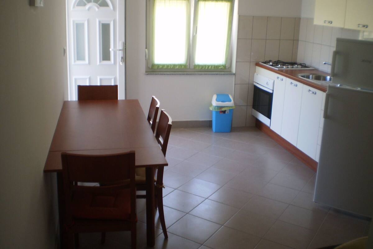 A kitchen with a dining table and chairs, a window with curtains, a trash bin, and a refrigerator.
