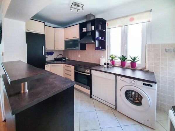 A compact kitchen with a built-in oven, a dishwasher, and a sink. There are cabinets with handles, a countertop, and a window with blinds. - Apartment Frano (Crikvenica)