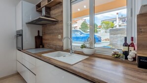 Moderni keittiö, jossa on puinen työtaso, valkoiset kaapit, sisäänrakennettu uuni ja ikkuna, josta avautuu näkymä siniseen autoon ja rakennuksiin.