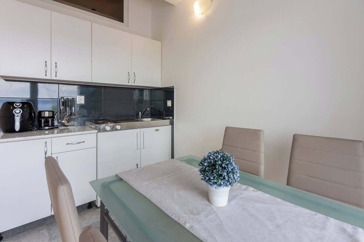 A compact kitchen with white cabinets, a coffee maker, and a sink. There is a table with a potted plant and a chair.