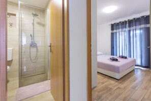 A hotel room with a shower, a bed with a pink bedspread, and a window with curtains. - Apartments Natalija (Novi Vinodolski)