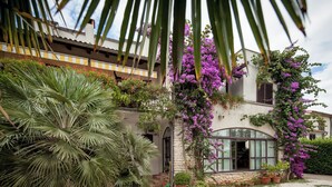 Uma casa com fachada de pedra, janelas amplas e um jardim com palmeiras e flores roxas.
