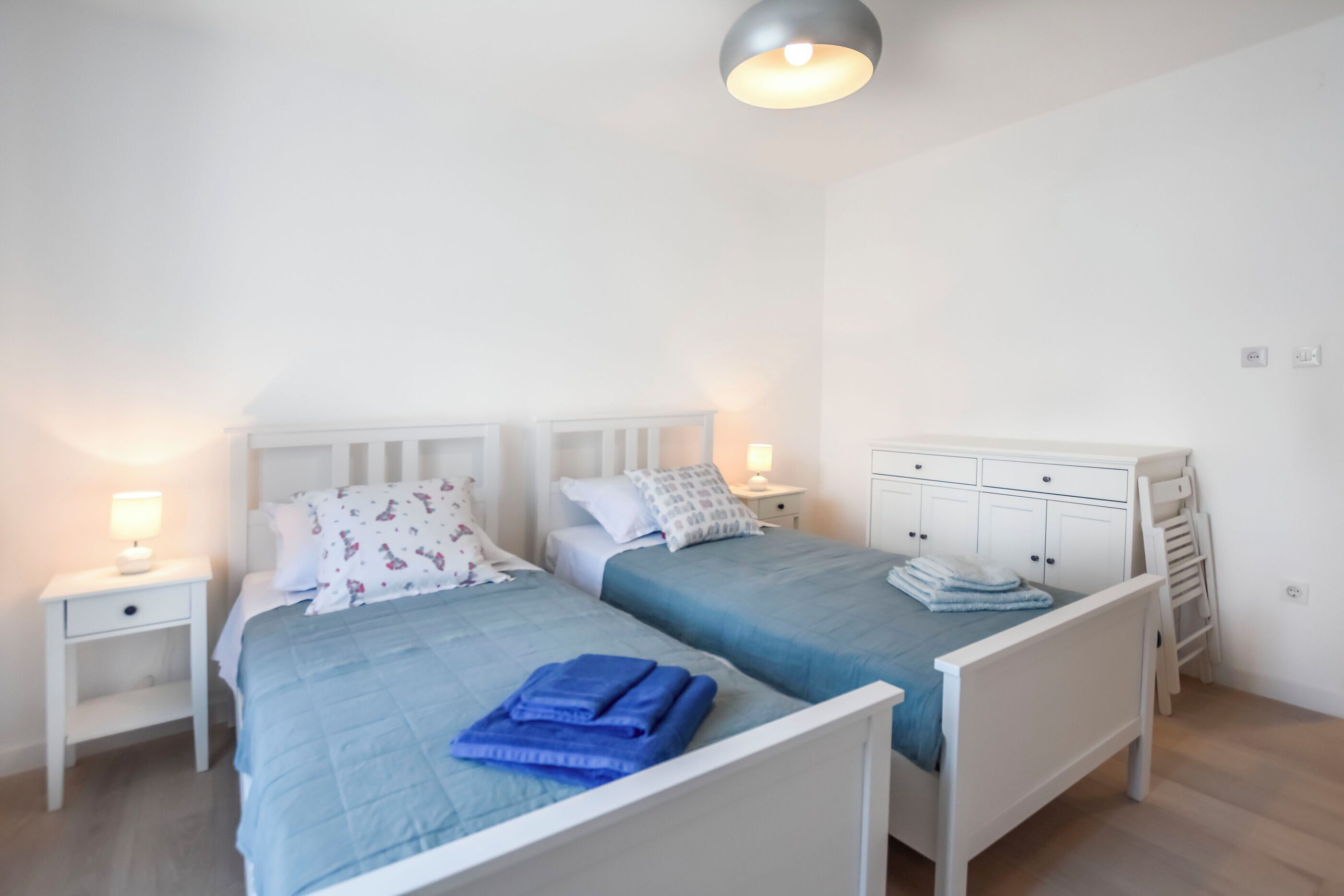 Une chambre avec deux lits, deux tables de chevet blanches, un luminaire au plafond et une armoire blanche.
