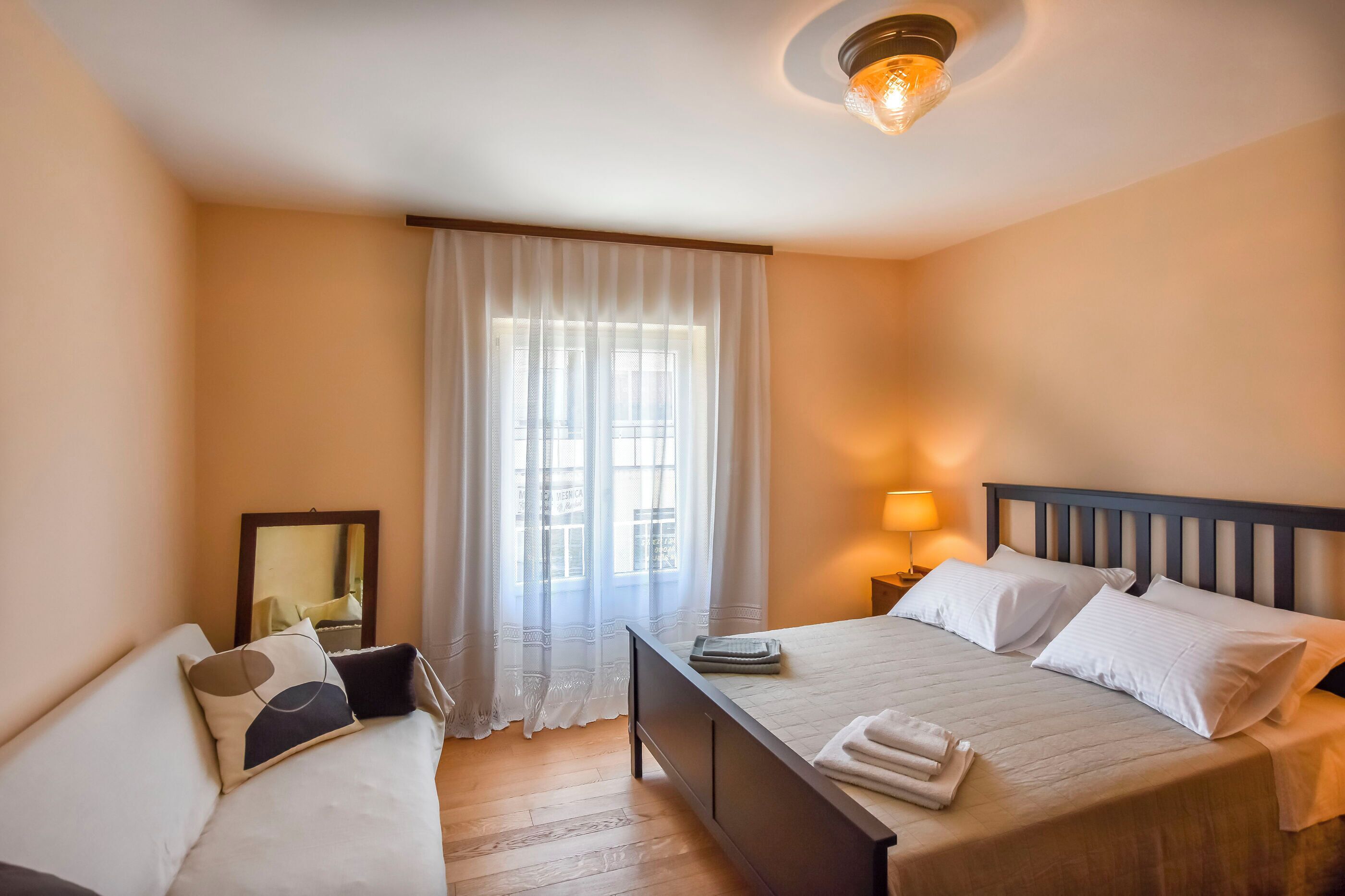 A hotel room with a bed, a bedside table, a lamp, a mirror, and a window with curtains.