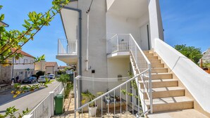 Un edificio moderno con facciata bianca, una scala che conduce a un balcone e vista sulla strada con auto parcheggiate e un albero.