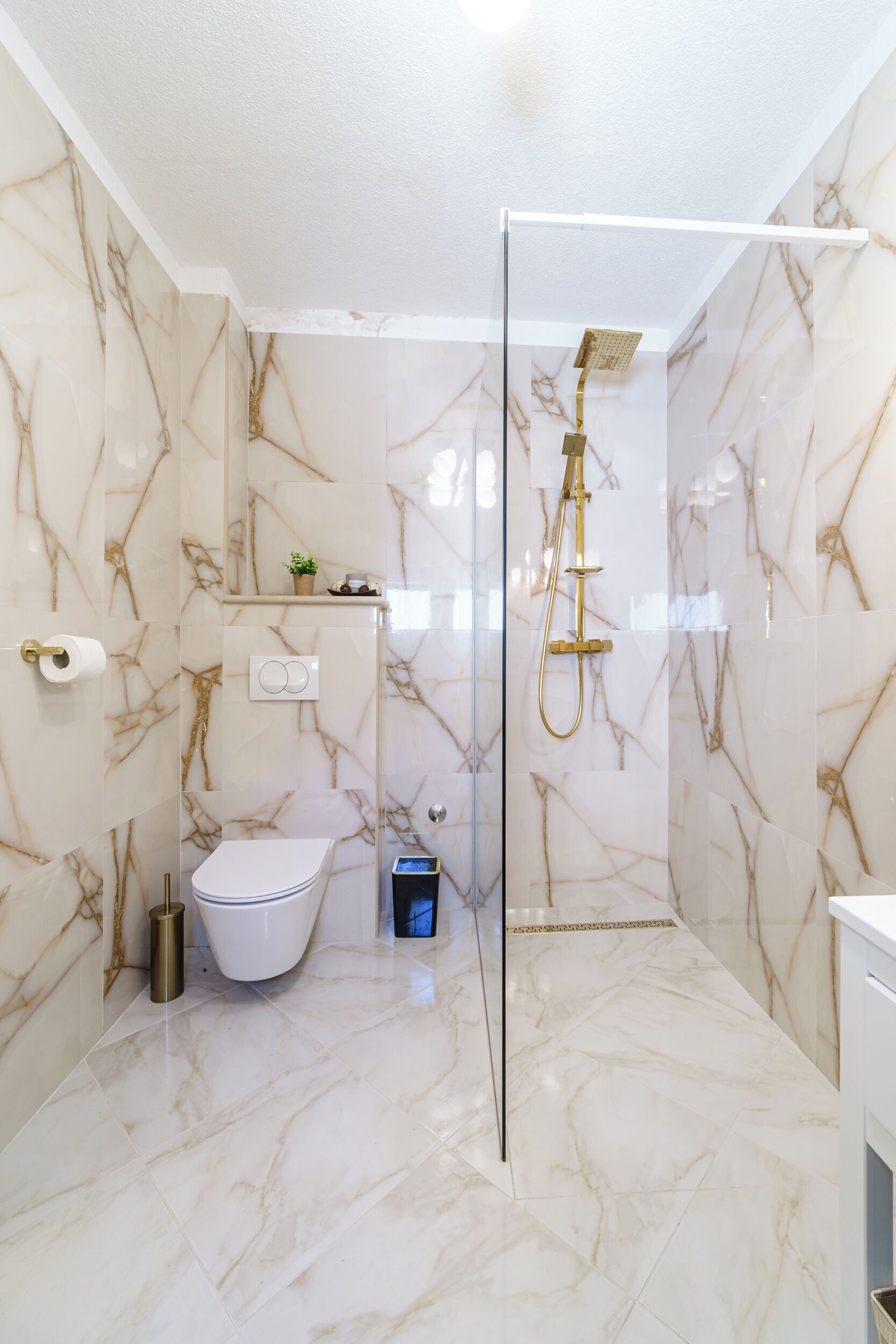 A bathroom with marble walls, a glass-enclosed shower, a toilet, and a small shelf with toiletries.