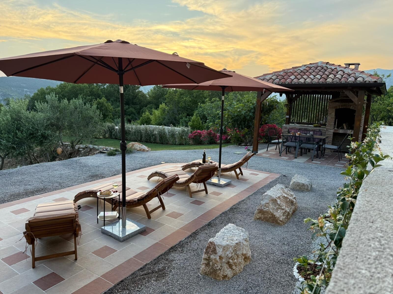 An outdoor patio with lounge chairs and a pergola, surrounded by a gravel path and greenery.